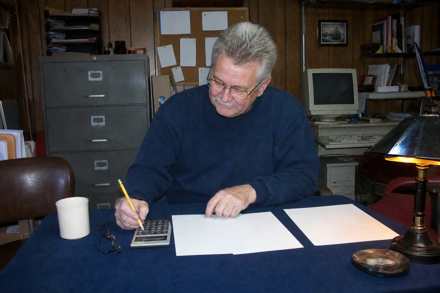An older man at a basement card table checks three blank quote sheets with a calculator.